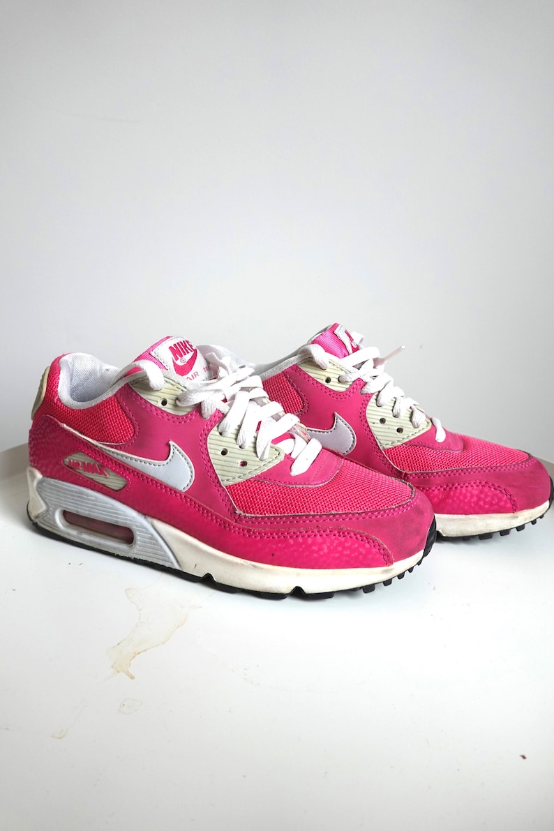K&ouml;nnte beinhalten: Paar pinkfarbene Nike Air Max 90 Sneaker. Die Schuhe haben wei&szlig;e Schn&uuml;rsenkel, eine wei&szlig;e und graue Sohle und einen wei&szlig;en Nike Swoosh. Die Zunge des Schuhs hat das Nike Air Logo. Die Schuhe stehen auf einer wei&szlig;en Oberfl&auml;che.