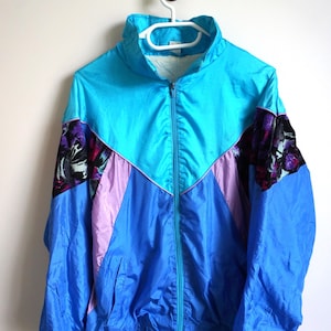 Può includere: Giacca a vento vintage blu, turchese e lavanda con chiusura a zip completa. La giacca presenta un design a blocchi di colore con un motivo floreale sulle maniche. È appesa a una gruccia bianca contro un muro bianco.