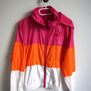 May include: A color-blocked jacket in pink, orange, and white, hanging on a white hanger. The jacket has a hood and a zipper closure. The brand "FILA" is visible on the chest. This athletic jacket is suitable for outdoor activities.