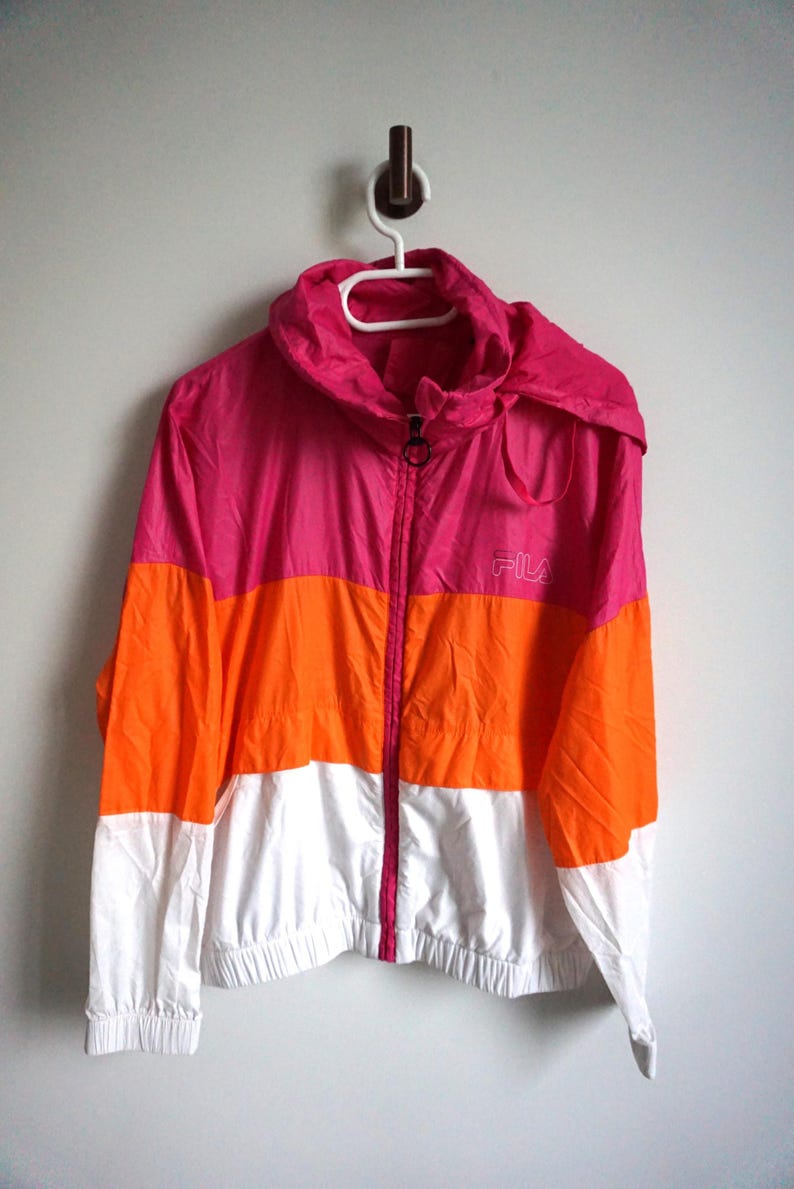May include: A color-blocked jacket in pink, orange, and white, hanging on a white hanger. The jacket has a hood and a zipper closure. The brand "FILA" is visible on the chest. This athletic jacket is suitable for outdoor activities.
