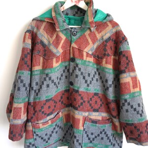 Könnte beinhalten: Eine Kapuzenjacke mit einem mehrfarbigen geometrischen Muster in Grau-, Rostorange- und Grüntönen. Das Kleidungsstück verfügt über eine Knopfleiste vorne, einen Kragen, lange Ärmel und zwei große aufgesetzte Taschen im unteren Vorderbereich.