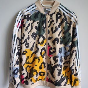 Puede incluir: Una chaqueta deportiva beige con cremallera y un estampado de leopardo multicolor en negro, amarillo, rosa y azul. La chaqueta incluye cuello, puños y dobladillo acanalados, con tres rayas blancas en cada manga y un logotipo de Adidas.
