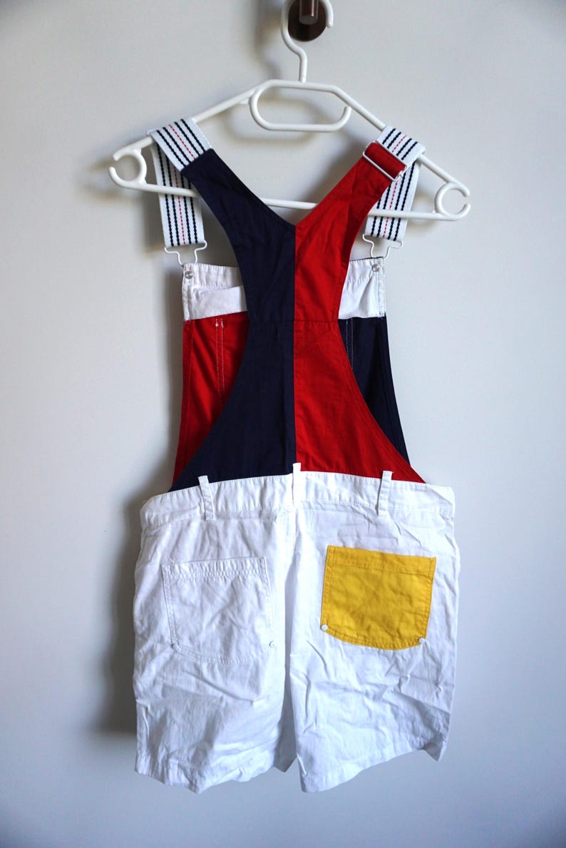 Puede incluir: Un pantal&oacute;n corto de peto blanco con un bolsillo amarillo, colgado de una percha blanca. La parte superior est&aacute; dividida verticalmente, con un lado azul marino y el otro rojo. Las correas son blancas con rayas azul marino y rojas.