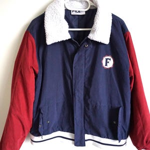 K&ouml;nnte beinhalten: Eine Vintage Fila Jacke in Marineblau mit roten &Auml;rmeln und einem wei&szlig;en, flauschigen Kragen. Die Jacke hat einen wei&szlig;-marinefarbenen gestreiften Saum und einen runden Aufn&auml;her mit dem Buchstaben "F".