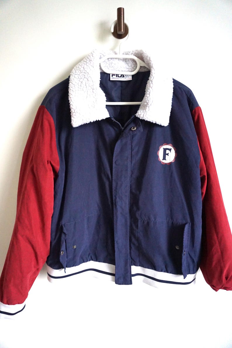 K&ouml;nnte beinhalten: Eine Vintage Fila Jacke in Marineblau mit roten &Auml;rmeln und einem wei&szlig;en, flauschigen Kragen. Die Jacke hat einen wei&szlig;-marinefarbenen gestreiften Saum und einen runden Aufn&auml;her mit dem Buchstaben "F".