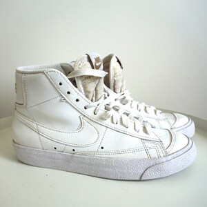 Op de afbeelding: Paar witte high-top sneakers met het Nike-logo op de zijkant en tong. De schoenen hebben een textuurzool en zijn geregen. De sneakers zijn schoon en zien er nieuw uit.