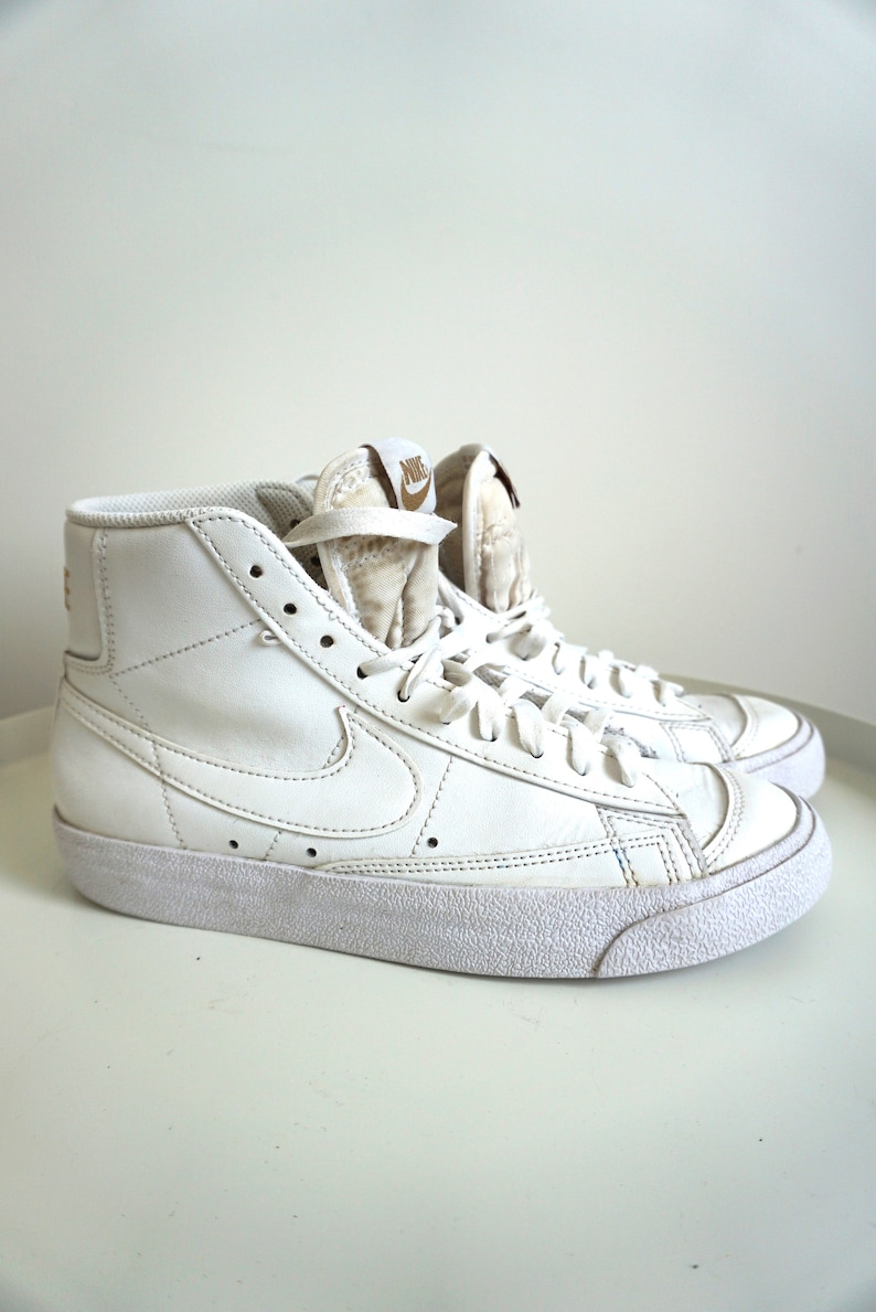 Op de afbeelding: Paar witte high-top sneakers met het Nike-logo op de zijkant en tong. De schoenen hebben een textuurzool en zijn geregen. De sneakers zijn schoon en zien er nieuw uit.