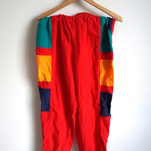 May include: Bright red vintage pants with color block accents in teal, yellow, and navy blue. The pants are hanging on a wooden hanger against a white wall. The elastic waistband is visible.