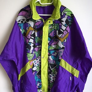 Può includere: Una giacca a vento con un vivace motivo geometrico in viola, verde lime, verde acqua e nero. La giacca presenta un colletto alto, un'abbottonatura centrale verde lime con bottoni a pressione, maniche lunghe e due tasche frontali con cerniera.