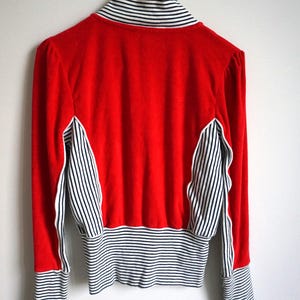May include: A red jacket with black and white striped accents on the sleeves, collar, and side panels. The jacket is hanging on a white wall. The jacket has long sleeves and a high collar.