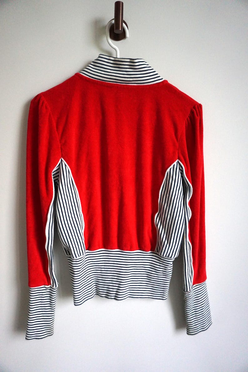 May include: A red jacket with black and white striped accents on the sleeves, collar, and side panels. The jacket is hanging on a white wall. The jacket has long sleeves and a high collar.