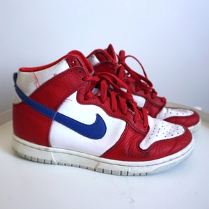 Puede incluir: Par de zapatillas altas rojas, blancas y azules. Las zapatillas presentan una parte superior de cuero rojo con paneles blancos y un logotipo azul de la marca. Cordones rojos y suela blanca.