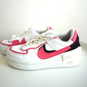 Puede incluir: Zapatillas blancas con detalles en rosa. Las zapatillas tienen una base blanca con detalles rosas en los laterales, el tal&oacute;n y la leng&uuml;eta. El ic&oacute;nico logo de Nike es de color rosa. La suela es blanca con la marca "AIR".