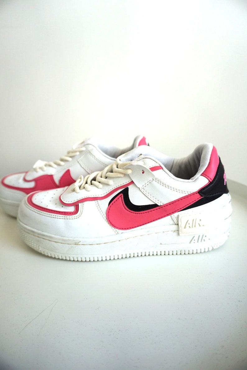 Puede incluir: Zapatillas blancas con detalles en rosa. Las zapatillas tienen una base blanca con detalles rosas en los laterales, el tal&oacute;n y la leng&uuml;eta. El ic&oacute;nico logo de Nike es de color rosa. La suela es blanca con la marca "AIR".