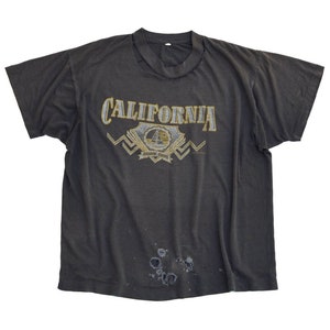 以下が含まれることがあります： 黄色の文字で「CALIFORNIA GOLDEN STATE」と印刷された、黒色のTシャツ。グラフィックには、帆船と太陽のスタイリッシュなイメージが描かれている。