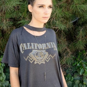以下が含まれることがあります： グレーのTシャツに「CALIFORNIA」の文字と金のグラフィックデザインが施された女性。彼女は長い銀色のネックレスにペンダントをつけています。