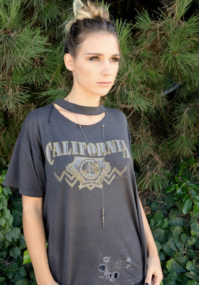 以下が含まれることがあります： グレーのTシャツに「CALIFORNIA」の文字と金のグラフィックデザインが施された女性。彼女は長い銀色のネックレスにペンダントをつけています。