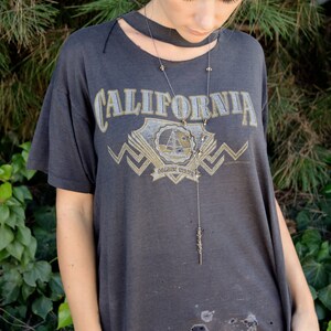 以下が含まれることがあります： 「CALIFORNIA GOLDEN STATE」とプリントされたグレーのTシャツを着た女性。彼女は長い細いネックレスにペンダントをつけている。
