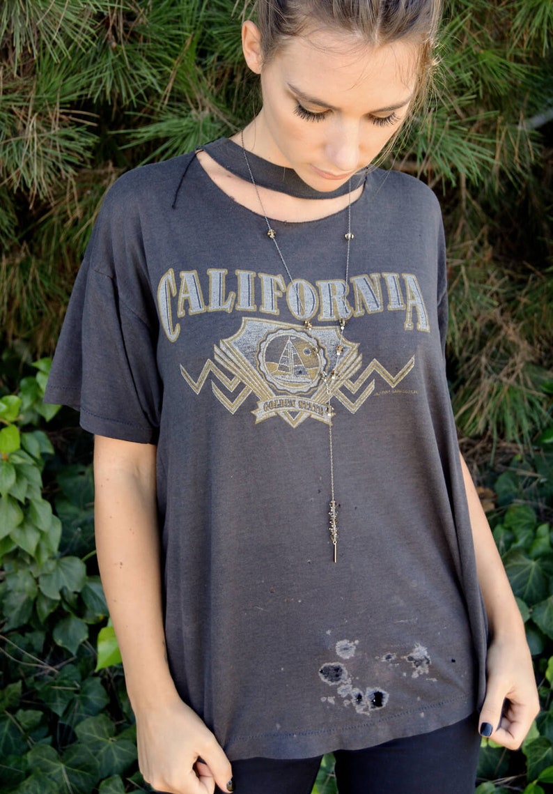 以下が含まれることがあります： 「CALIFORNIA GOLDEN STATE」とプリントされたグレーのTシャツを着た女性。彼女は長い細いネックレスにペンダントをつけている。