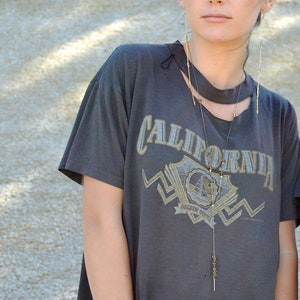 以下が含まれることがあります： 「CALIFORNIA」とプリントされたグレーのTシャツを着た女性。彼女は小さなペンダントの付いた長いゴールドネックレスと長いゴールドのイヤリングをしています。
