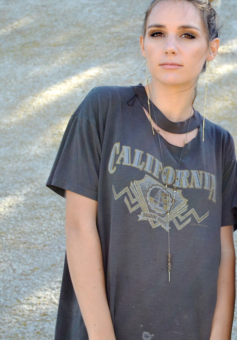 以下が含まれることがあります： 「CALIFORNIA」とプリントされたグレーのTシャツを着た女性。彼女は小さなペンダントの付いた長いゴールドネックレスと長いゴールドのイヤリングをしています。