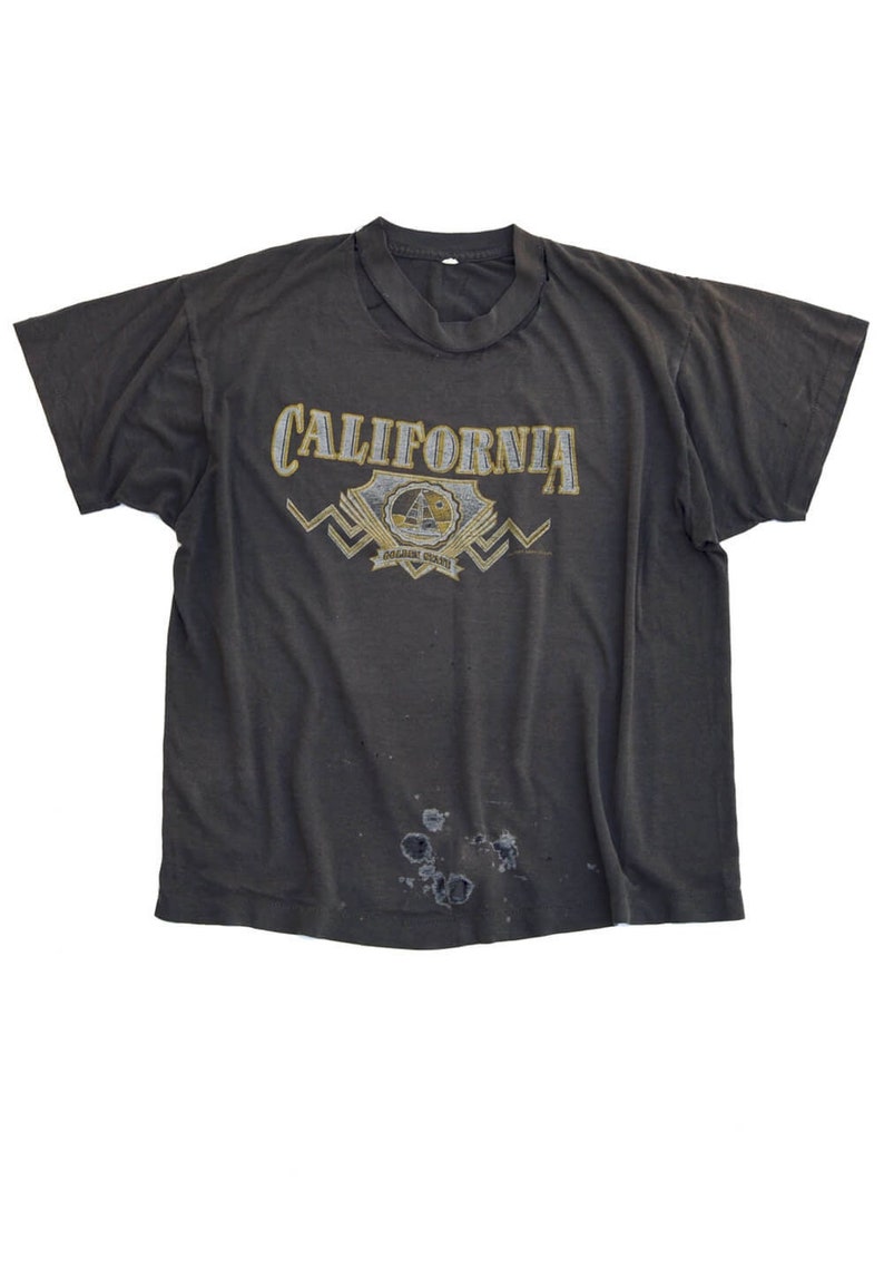 以下が含まれることがあります： 黄色の文字で「CALIFORNIA GOLDEN STATE」と印刷された、黒色のTシャツ。グラフィックには、帆船と太陽のスタイリッシュなイメージが描かれている。