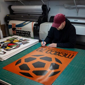 May include: A rectangular poster featuring a large black soccer ball graphic on an orange background with the text MEXICO 70 and IX campeonato mundial de futbol, mayo 31 - junio 21 printed in black ink on the lower portion of the paper sheet.