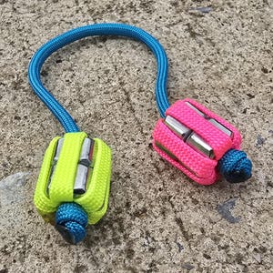 May include: Two colorful paracord accessories. One is neon yellow, the other is hot pink. Both have silver metal inserts and are connected by a blue paracord loop. The accessories are knotted at the ends.