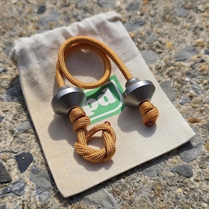 May include: A yellow and silver fidget toy with a knotted rope design. The toy has two silver metal ends and a yellow rope connecting them. It rests on a beige cloth bag with a green logo.