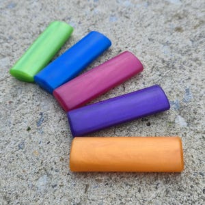May include: A collection of six colorful, rectangular wooden beads in shades of green, blue, pink, purple, and orange. The beads are arranged on a rough, gray concrete surface, showcasing their vibrant colors and smooth texture.
