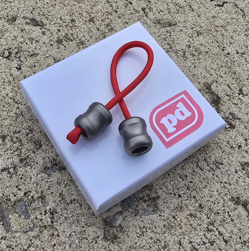 K&ouml;nnte beinhalten: Ein rotes Schl&uuml;sselband mit zwei grauen, fassf&ouml;rmigen Perlen. Das Schl&uuml;sselband liegt auf einer wei&szlig;en Schachtel mit einem rosa Logo mit der Aufschrift "pd". Der Hintergrund ist eine strukturierte, graue Oberfl&auml;che.