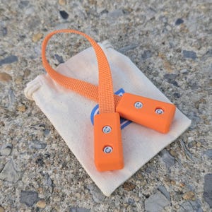 May include: An orange fabric strap with two rectangular orange blocks attached, each featuring two silver metal rivets. The strap is positioned over a small, off-white cotton drawstring pouch that displays a partial blue logo print.