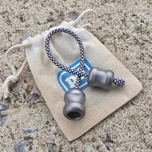 May include: A gray metal begleri set with a black and white patterned cord, on a small beige drawstring bag. The bag features a blue logo with a white "P". The begleri has a cylindrical shape.