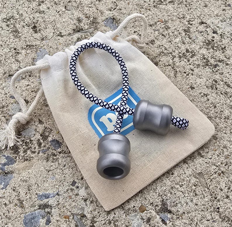 K&ouml;nnte beinhalten: Ein graues Metall-Begleri-Set mit einem schwarz-wei&szlig; gemusterten Kordel, das auf einem kleinen beigen Kordelzugbeutel liegt. Die Tasche hat ein blaues Logo mit einem wei&szlig;en "P". Das Begleri hat eine zylindrische Form.