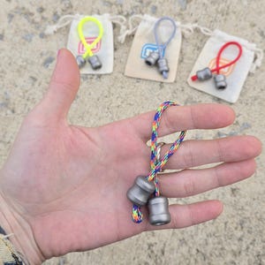 K&ouml;nnte beinhalten: Nahaufnahme eines kleinen, silberfarbenen Fidget-Spielzeugs mit einer regenbogenfarbenen Schnur. Das Spielzeug wird in einer Hand gehalten, mit drei weiteren &auml;hnlichen Spielzeugen im Hintergrund, die jeweils an einem kleinen Kordelzugbeutel mit einer farbigen Schlaufe befestigt sind.