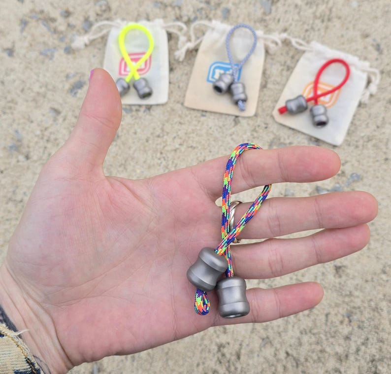 K&ouml;nnte beinhalten: Nahaufnahme eines kleinen, silberfarbenen Fidget-Spielzeugs mit einer regenbogenfarbenen Schnur. Das Spielzeug wird in einer Hand gehalten, mit drei weiteren &auml;hnlichen Spielzeugen im Hintergrund, die jeweils an einem kleinen Kordelzugbeutel mit einer farbigen Schlaufe befestigt sind.