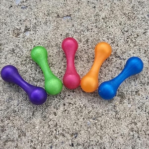 Op de afbeelding: Vijf kleurrijke dumbbell-vormige hondenspeeltjes in paars, groen, roze, oranje en blauw. Elk speeltje heeft een afgeronde vorm aan beide uiteinden, verbonden door een smaller midden. De speeltjes zijn gerangschikt op een getextureerd, grijs oppervlak.