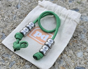 Puede incluir: Un cordón de paracord verde con dos juegos de tres cuentas metálicas cilíndricas de bordes redondeados, aseguradas con nudos, sobre una pequeña bolsa de tela color crema con un logotipo cuadrado naranja con las letras pd.