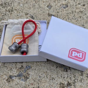 K&ouml;nnte beinhalten: Zwei graue Metallperlen, verbunden durch eine rote Schnur, in einer offenen wei&szlig;en Schachtel mit pinkfarbenem Logo. Ein kleiner beigefarbener Beutel mit Kordelzug ist ebenfalls zu sehen. Die Perlen haben eine abgerundete Form.