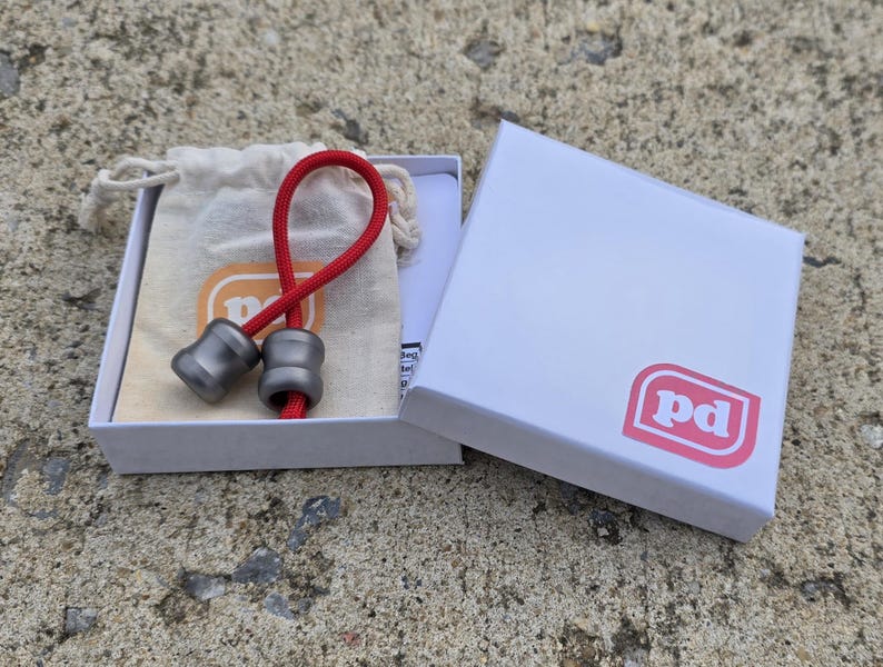 K&ouml;nnte beinhalten: Zwei graue Metallperlen, verbunden durch eine rote Schnur, in einer offenen wei&szlig;en Schachtel mit pinkfarbenem Logo. Ein kleiner beigefarbener Beutel mit Kordelzug ist ebenfalls zu sehen. Die Perlen haben eine abgerundete Form.