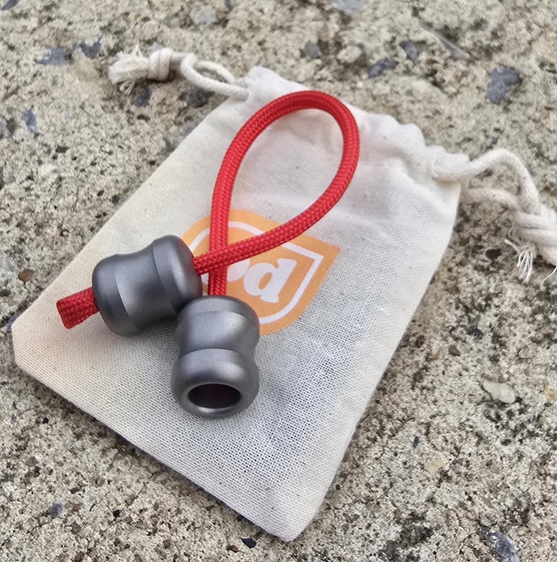 K&ouml;nnte beinhalten: Zwei graue Metallperlen, verbunden durch eine rote Kordel, liegen auf einem kleinen, cremefarbenen Beutel mit Kordelzug. Der Beutel hat ein Logo mit dem Buchstaben "d". Die Perlen haben eine einzigartige, sanduhrf&ouml;rmige Gestalt. Der Hintergrund ist eine strukturierte, helle Oberfl&auml;che.