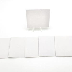 May include: A collection of white, square-shaped cards, some laid flat and others displayed upright on a clear plastic stand. The cards have a smooth, blank surface, suitable for various uses.