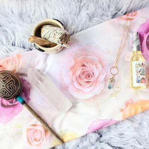 May include: A white floral fabric with a wooden maraca, a clear quartz crystal, a small white ceramic bowl with a sage smudge stick, a gold chain necklace with a green crystal pendant, and a small glass bottle of oil.