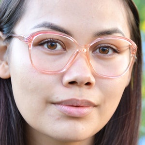 May include: A close-up of a person wearing pink, plastic-framed glasses. The glasses have a rounded, retro style.