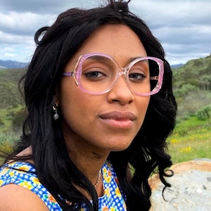 May include: A person wearing large, clear-framed spectacles with pink rims and gold accents. The spectacles have a square shape with rounded edges. The person has dark hair and is wearing a blue and yellow floral patterned top.