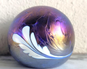 May include: A decorative glass sphere with a metallic purple and gold finish. A white and blue floral design adorns the side of the sphere. The sphere is sitting on a marble surface.