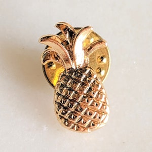 May include: A gold-toned pineapple pin with a detailed design. The pin is attached to a circular backing with a safety clasp.