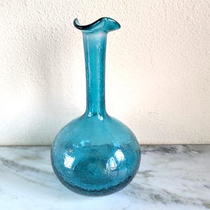 May include: A teal-colored glass vase with a round base and a long, slender neck. The vase has a textured, crackled appearance and a flared, ruffled rim. The vase is displayed on a marble surface.