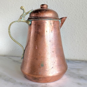 Puede incluir: Una tetera de cobre vintage con tapa y asa de latón. La tetera tiene una forma clásica con un pico para verter. El cobre tiene un color marrón rojizo cálido con algunas áreas de pátina, lo que le da una apariencia rústica.