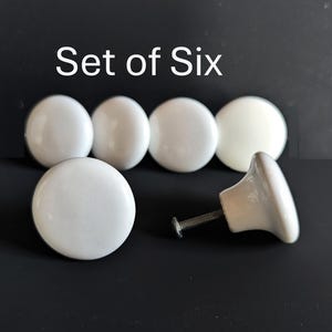 May include: Set of six white ceramic cabinet knobs. The knobs are round with a glossy finish. One knob is shown with its screw attached. The text "Set of Six" is above the knobs.